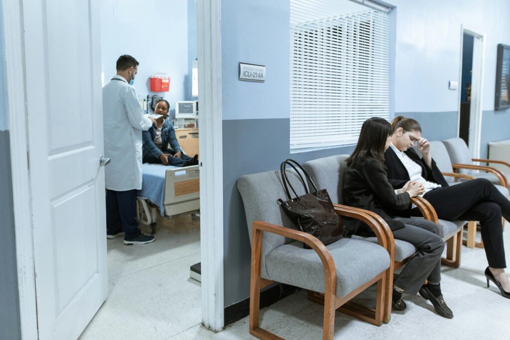 Doctor with patient in ICU, women in waiting area showing concern.